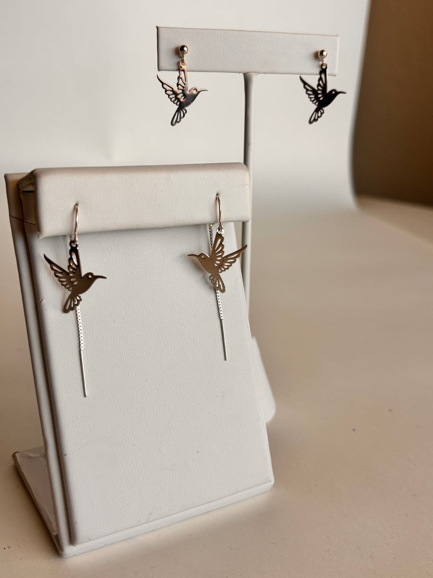 Two styles of silver hummingbird earrings displayed on white jewelry stands against a neutral background. The top stand showcases hummingbird charm stud earrings, while the front stand features threader-style earrings with dangling hummingbird cutouts and fine chain detail, emphasizing their delicate, airy design.