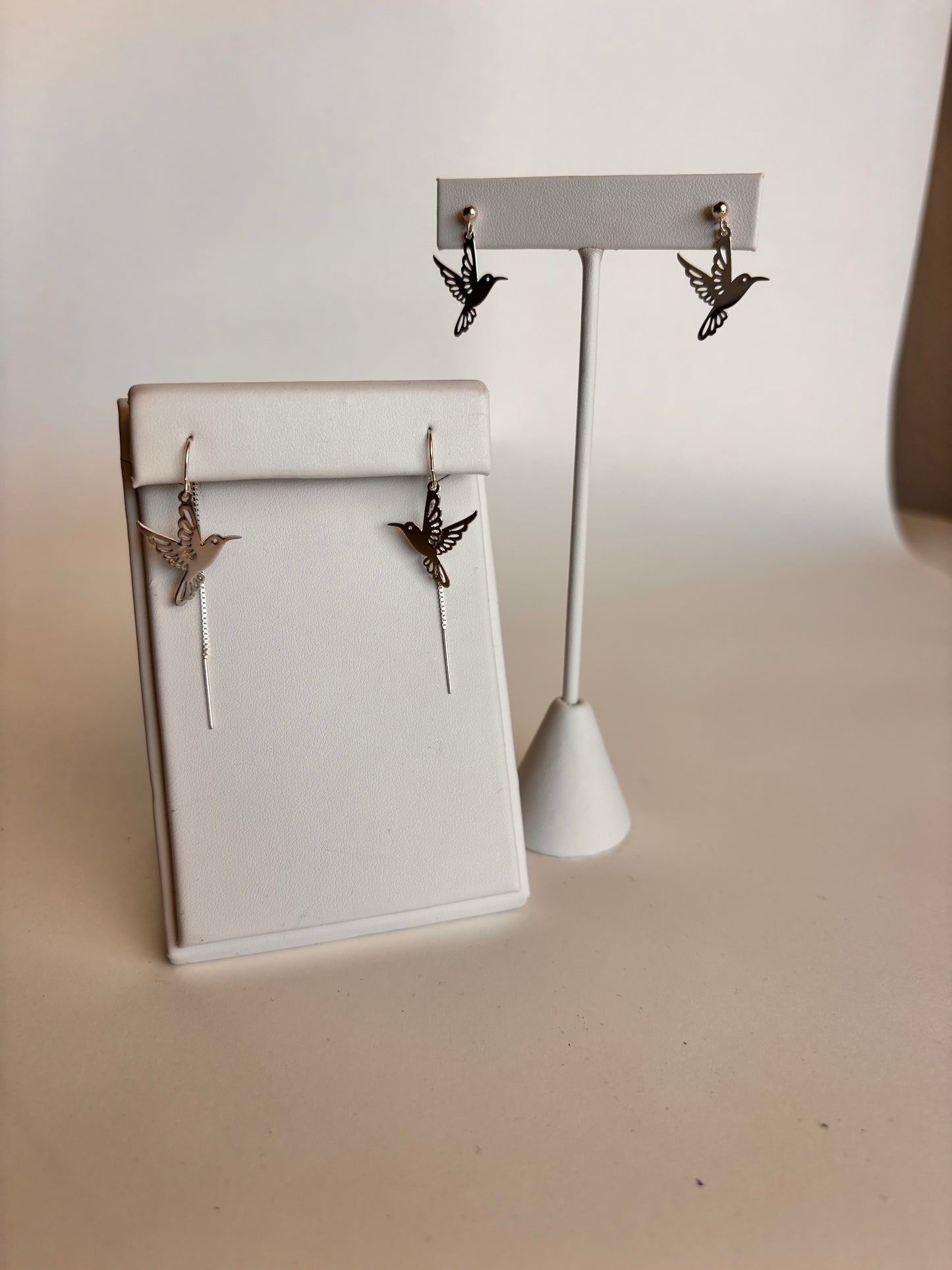 Two pairs of silver hummingbird earrings displayed on white jewelry stands against a soft neutral background. The front stand features threader-style earrings with dangling cutout hummingbirds and fine chain detail, while the back stand displays matching hummingbird stud earrings for a coordinated yet varied look.