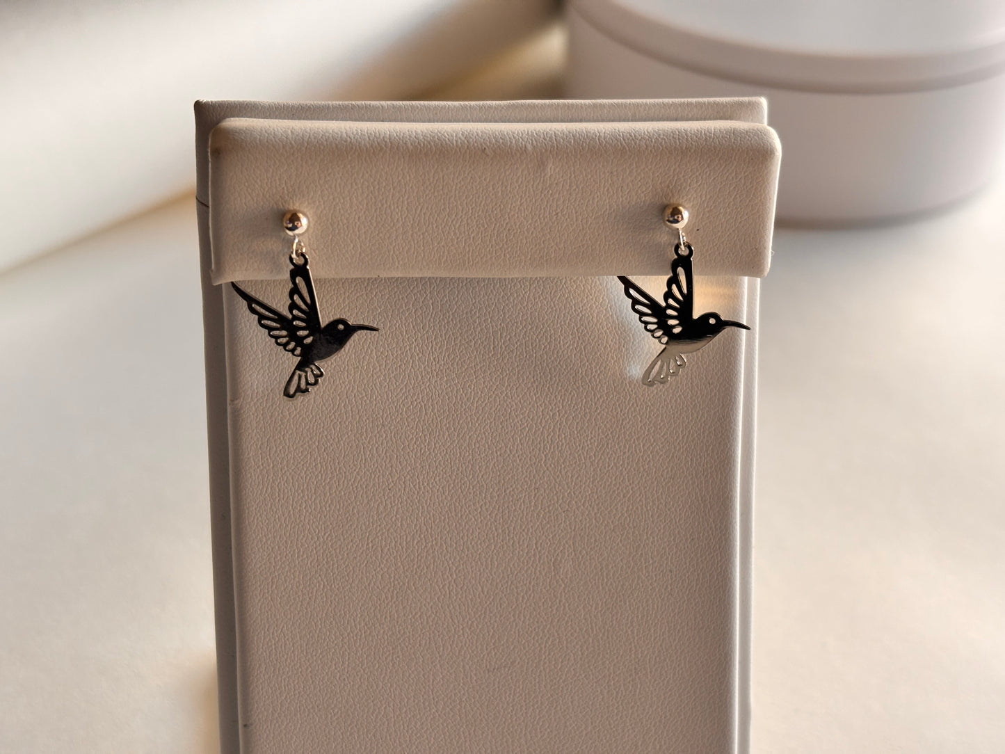 Delicate black hummingbird silhouette earrings displayed on a cream-colored jewelry stand with a soft, neutral background featuring a round white box and warm natural lighting.