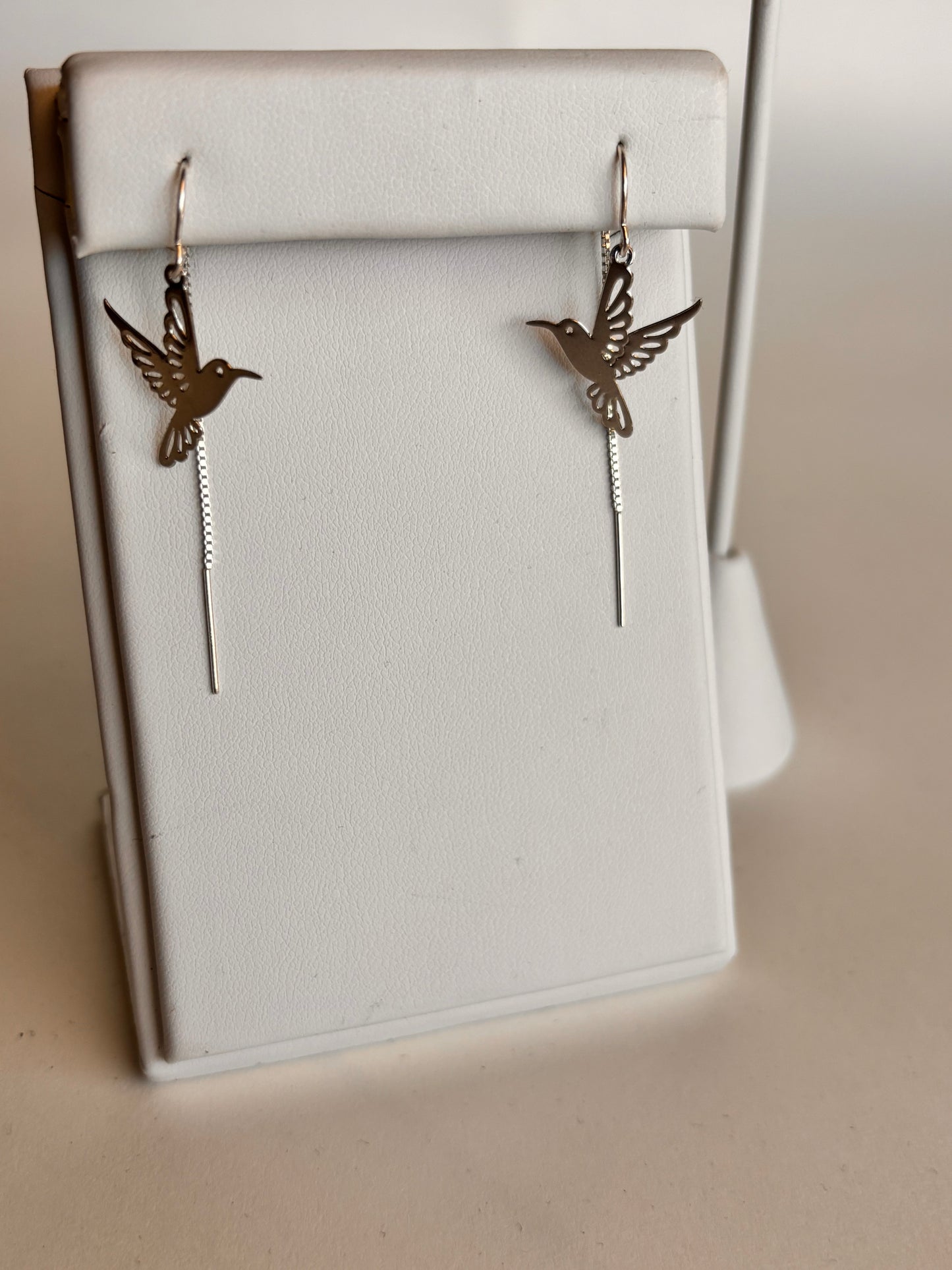 Close-up of a pair of silver threader earrings featuring delicate cutout hummingbird charms, displayed on a white jewelry stand against a neutral background. The fine chain detail and minimalist design create an airy, lightweight look.